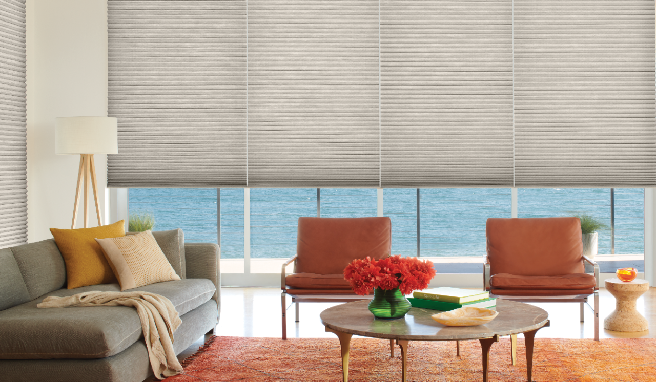 Large, brightly lit living room featuring ceiling to floor windows with shades behind a sofa and accent chair. 